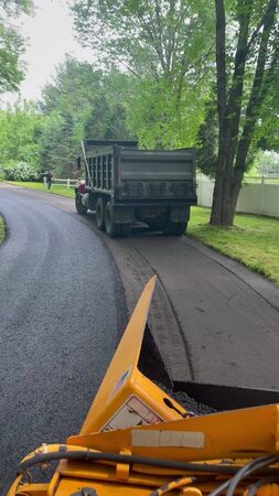 An asphalt paving machine with its hopper full of dark hot-mix asphalt follows a dump truck up a curving residential driveway lined by green trees and a white fence.