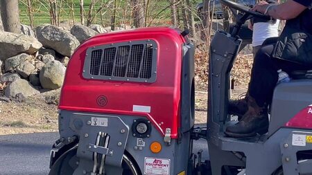 Close-up of a red and gray asphalt roller being driven along a section of new blacktop, with a stone wall and bare trees in the background.