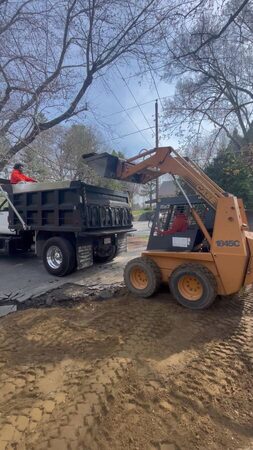 A CASE 1845C skid-steer loader lifts its bucket beside a black dump truck being loaded with old asphalt and debris from a driveway demolition; a worker in an orange jacket sits in the truck bed.