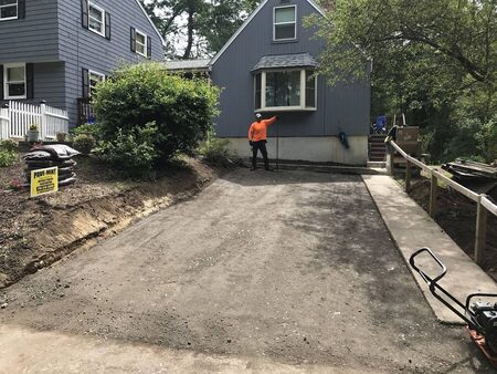 A crew member in an orange high-visibility shirt stands at a graded gravel driveway base holding a stake, beside a yellow PAVE-MINT yard sign and bagged mulch, prepping for paving next to a gray house.