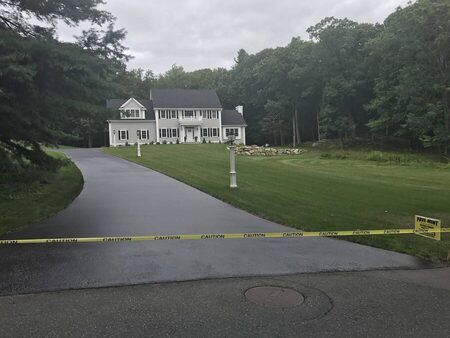 Curved, freshly sealcoated driveway leading to a large white colonial-style home with black shutters, bordered by manicured lawn and a stone wall; yellow CAUTION tape and a PAVE-MINT yard sign block the driveway entrance.