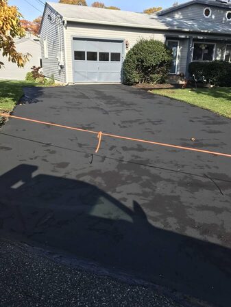 A freshly sealcoated driveway with a mottled, drying marbled texture and an orange barrier line strung across it, leading to a single-car garage in autumn foliage.