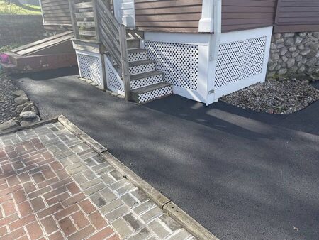 Newly paved asphalt walkway curving beside a brick paver patio and wooden deck stairs with white lattice skirting, running along the foundation of a house.