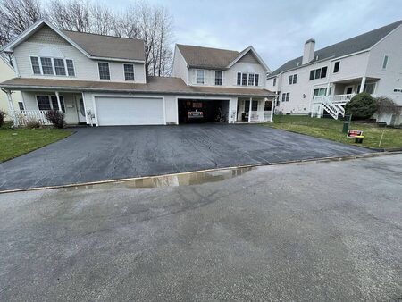 Completed black asphalt driveway of a two-car-garage house under overcast sky, still glossy with fresh sealcoat, clean straight edging along the lawn, and a PAVE-MINT yard sign visible near the street.
