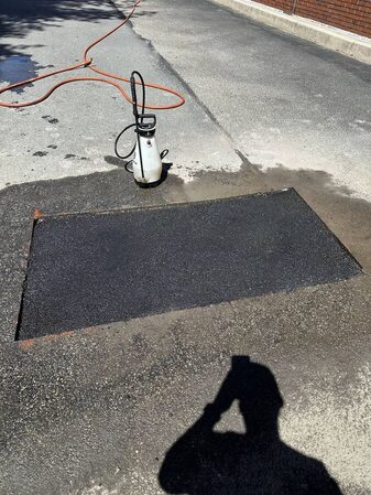 Overhead close-up of a fresh rectangular asphalt patch repair near a brick building, with an orange hose and pump sprayer nearby.