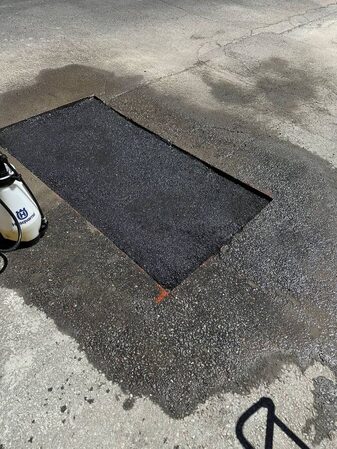 Overhead close-up of a rectangular fresh asphalt patch repair set into surrounding older pavement, with a white backpack sprayer resting beside it.