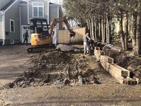 A CASE mini-excavator and two crew members work on a segmental block retaining wall along a driveway excavation beside a gray house; gravel base material and a partially built wall are visible.