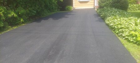 A freshly paved straight driveway bordered by lush hosta plantings and evergreen shrubs, leading to a yellow house garage.