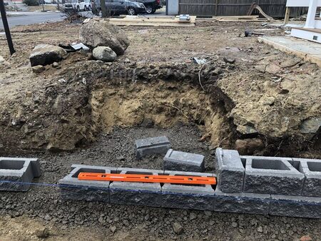 Close-up of gray segmental retaining wall block being set and leveled with an orange line level along a blue chalk line, on a gravel base beside an excavated pit with large boulders.