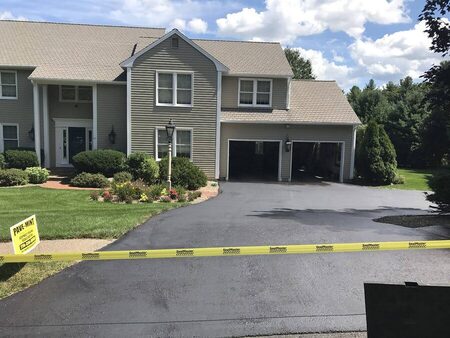 Freshly sealcoated driveway of a two-story tan colonial home with a two-car garage, landscaped front beds in bloom, a yellow PAVE-MINT yard sign, and yellow SealMaster barrier tape strung across the driveway.