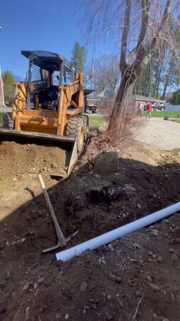 An orange skid-steer loader with bucket lowered into a dirt driveway excavation trench, a white drainage pipe and pickaxe on the ground, two crew members visible in the background near a paved area.