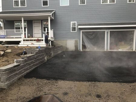 Steam rising off a freshly laid section of hot asphalt driveway next to a completed gray segmental block retaining wall; a two-car garage with doors covered in protective plastic sheeting stands in the background, a crew member watches from the wall.