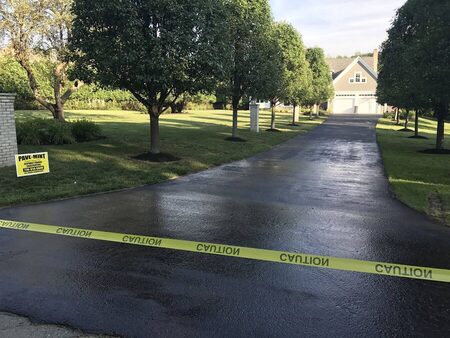 Sunlit view down a long, freshly sealcoated driveway lined with matching ornamental trees between brick entry pillars, a yellow PAVE-MINT yard sign and caution tape in the foreground, leading up to a large tan house with a two-car garage.