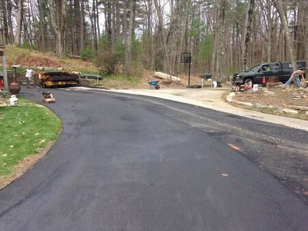 A newly paved wooded driveway curving past a yellow asphalt paving machine and an orange vibratory plate compactor parked at the top, with a basketball hoop, wheelbarrow, and crew pickup truck near a paved turnaround.