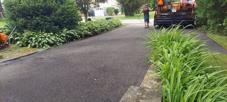 A worker hoses off a yellow asphalt paving machine parked on a freshly paved driveway bordered by hostas, boxwood hedges, and ornamental grasses.