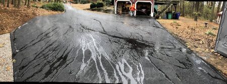 Wide panoramic shot of a worker in an orange hoodie spray-applying sealcoat with a hose along a long, tree-lined driveway; the wet sealcoat shows dramatic dark streaking and pooling texture, garage and parked car visible ahead.
